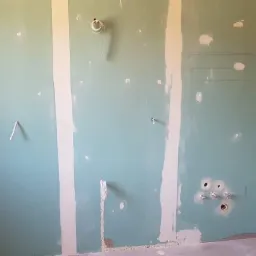 Ściana z płyt kartonowo-gipsowych w trakcie montażu, widoczne przewody elektryczne i rury, szpachlowane łączenia płyt.