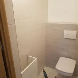 Montaż szafki pod umywalkę w łazience z wiszącą toaletą, widoczna wiertarka i narzędzia na podłodze.