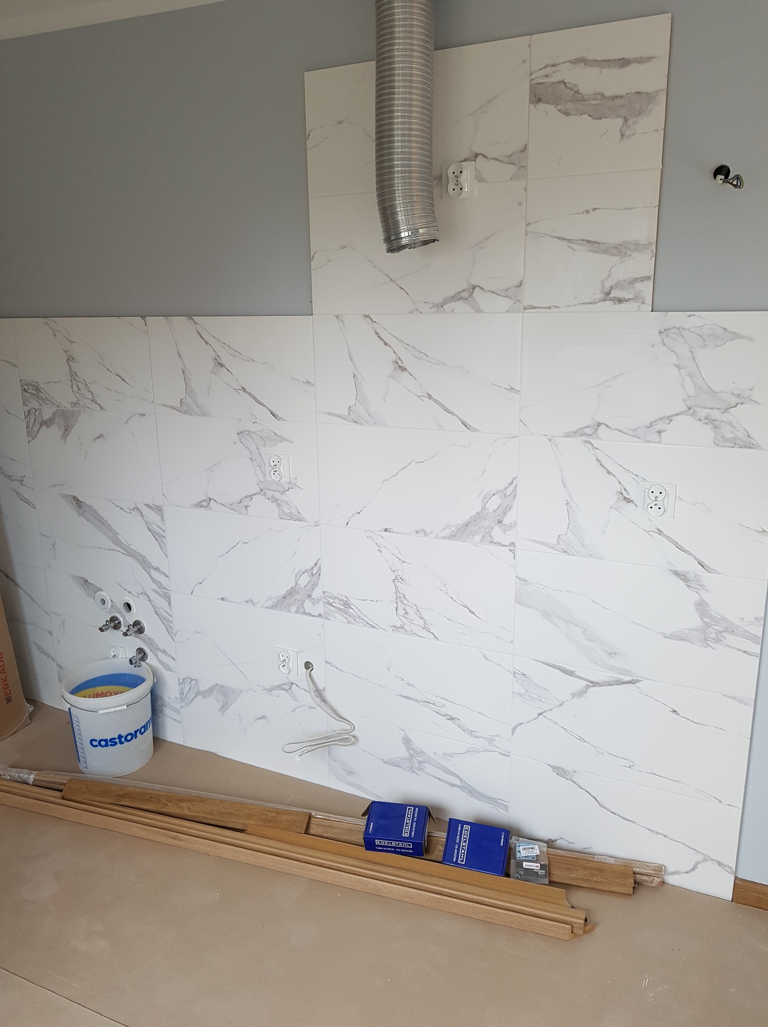 Ściana w trakcie wykańczania z białych płytek imitujących marmur, widoczne puszki elektryczne, rury, wiadro z farbą Castorama, listwy przypodłogowe i niebieskie opakowania.