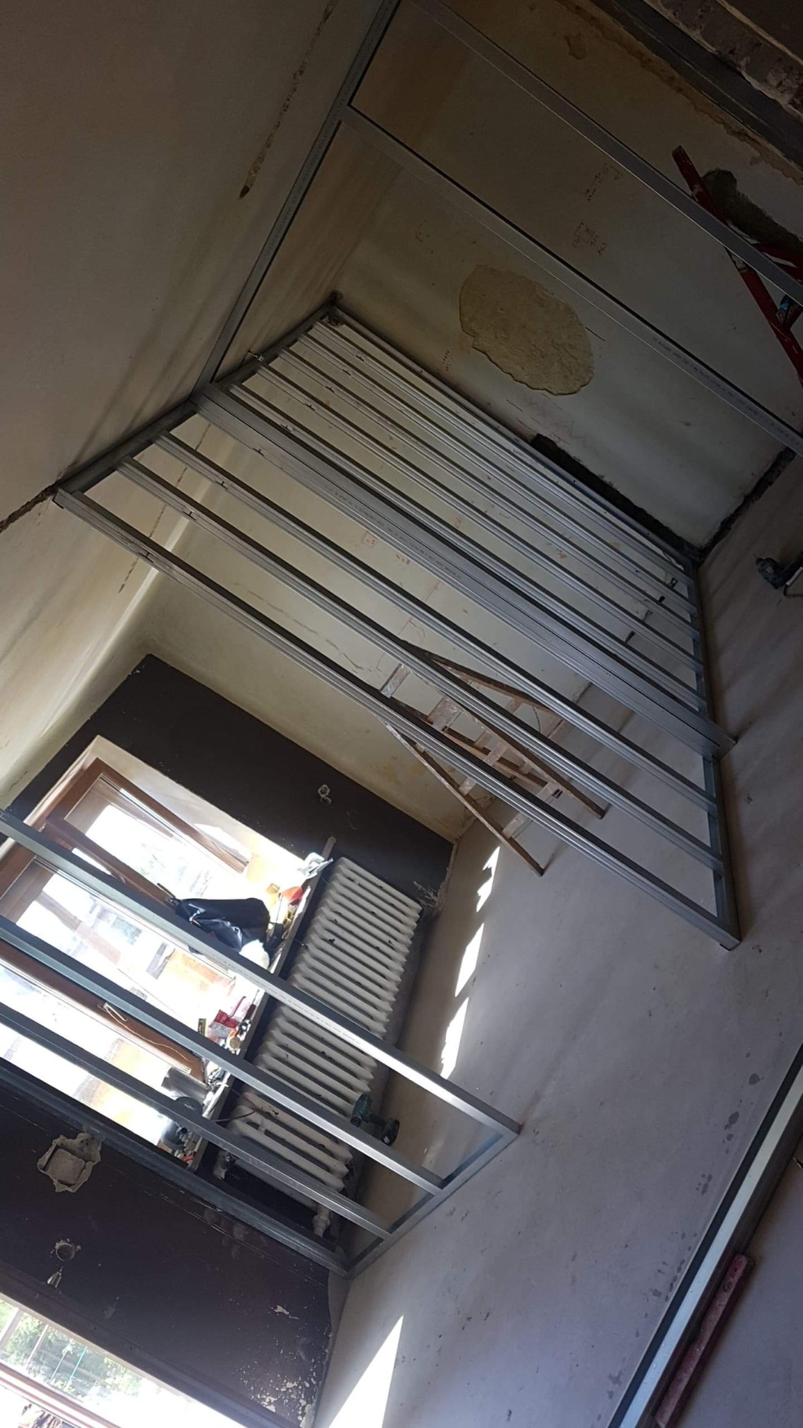 Montaż metalowego stelażu pod płyty karton gips na ścianie, widoczne profile, okno z grzejnikiem i fragment drabiny.