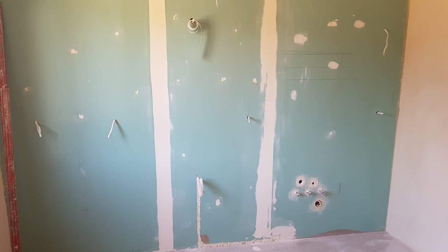 Ściana z płyt kartonowo-gipsowych w trakcie montażu, widoczne przewody elektryczne i rury, szpachlowane łączenia płyt.