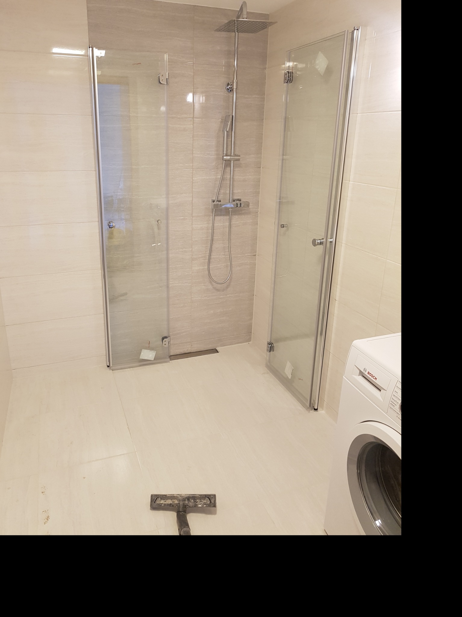Nowoczesna łazienka z kabiną prysznicową typu walk-in, wykończona beżowymi płytkami, z widoczną pralką marki Bosch i narzędziem do fugowania na podłodze.