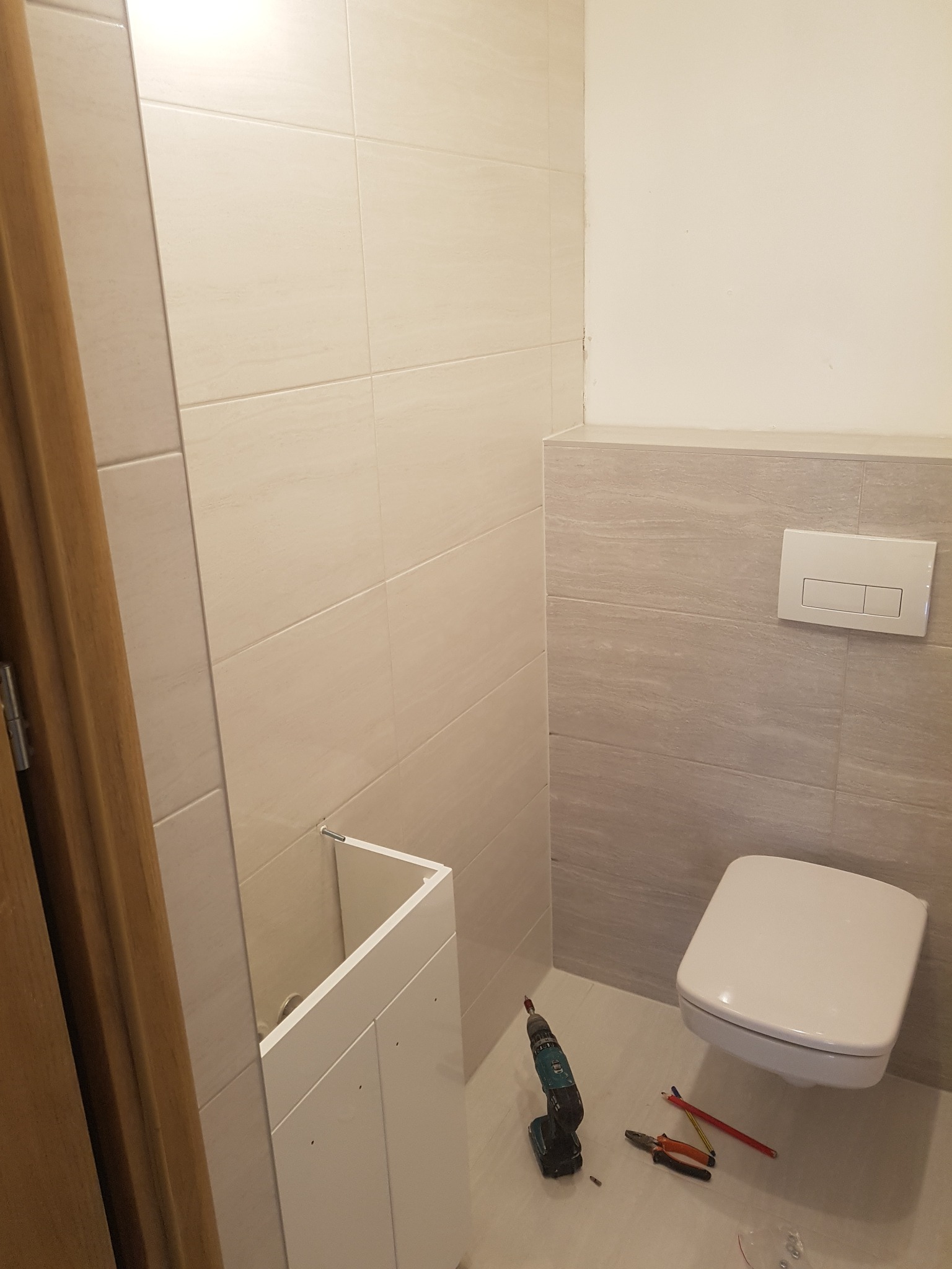 Montaż szafki pod umywalkę w łazience z wiszącą toaletą, widoczna wiertarka i narzędzia na podłodze.