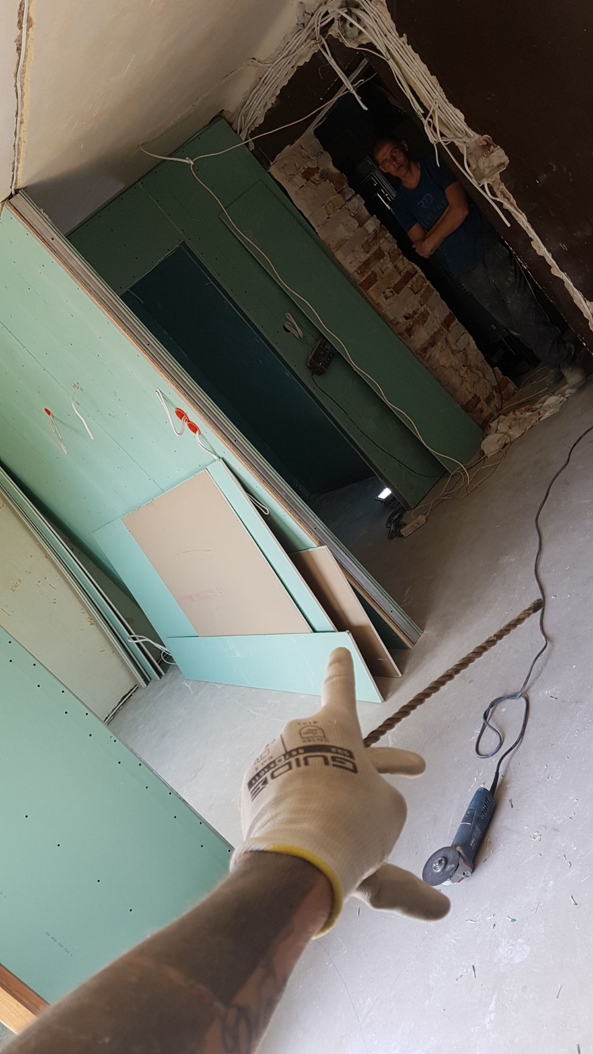 Montaż płyt karton-gips w Kielcach: widok z perspektywy osoby wskazującej na ułożone płyty, w tle widoczna osoba w przejściu i narzędzie szlifierskie na podłodze.