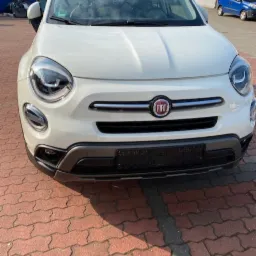 Biały samochód marki Fiat, model 500X, zaparkowany na czerwonej kostce brukowej, z widocznym przodem pojazdu i fragmentem otoczenia warsztatowego.