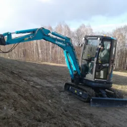 Niebieska mini koparka Sunward SWE25 pracuje na zboczu ziemnym, z operatorem w kabinie, w tle drzewa bez liści i fragment ogrodzenia.