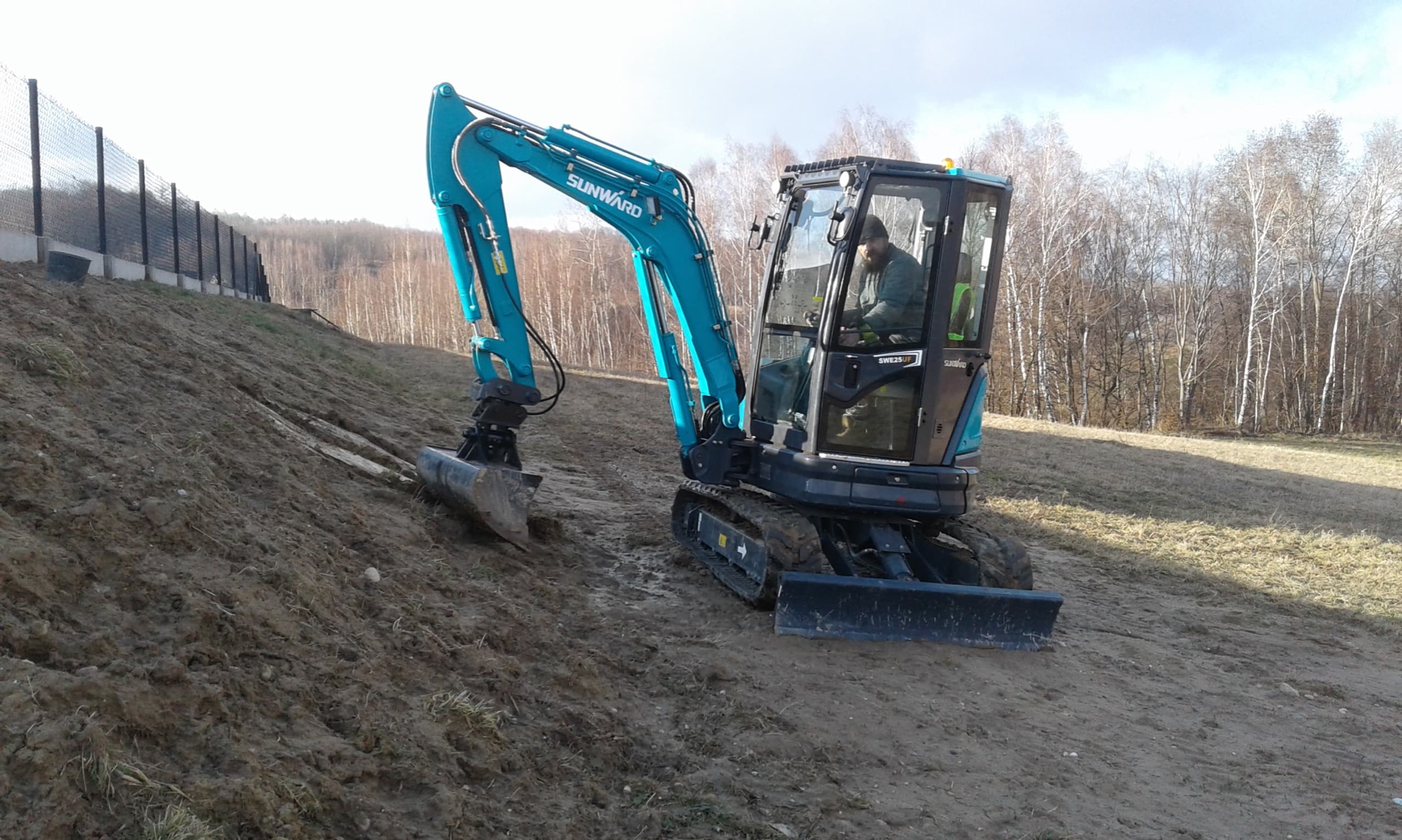 Niebieska mini koparka Sunward SWE25UF z operatorem, pracująca na zboczu z widocznymi śladami gąsienic, w tle ogrodzenie i drzewa.