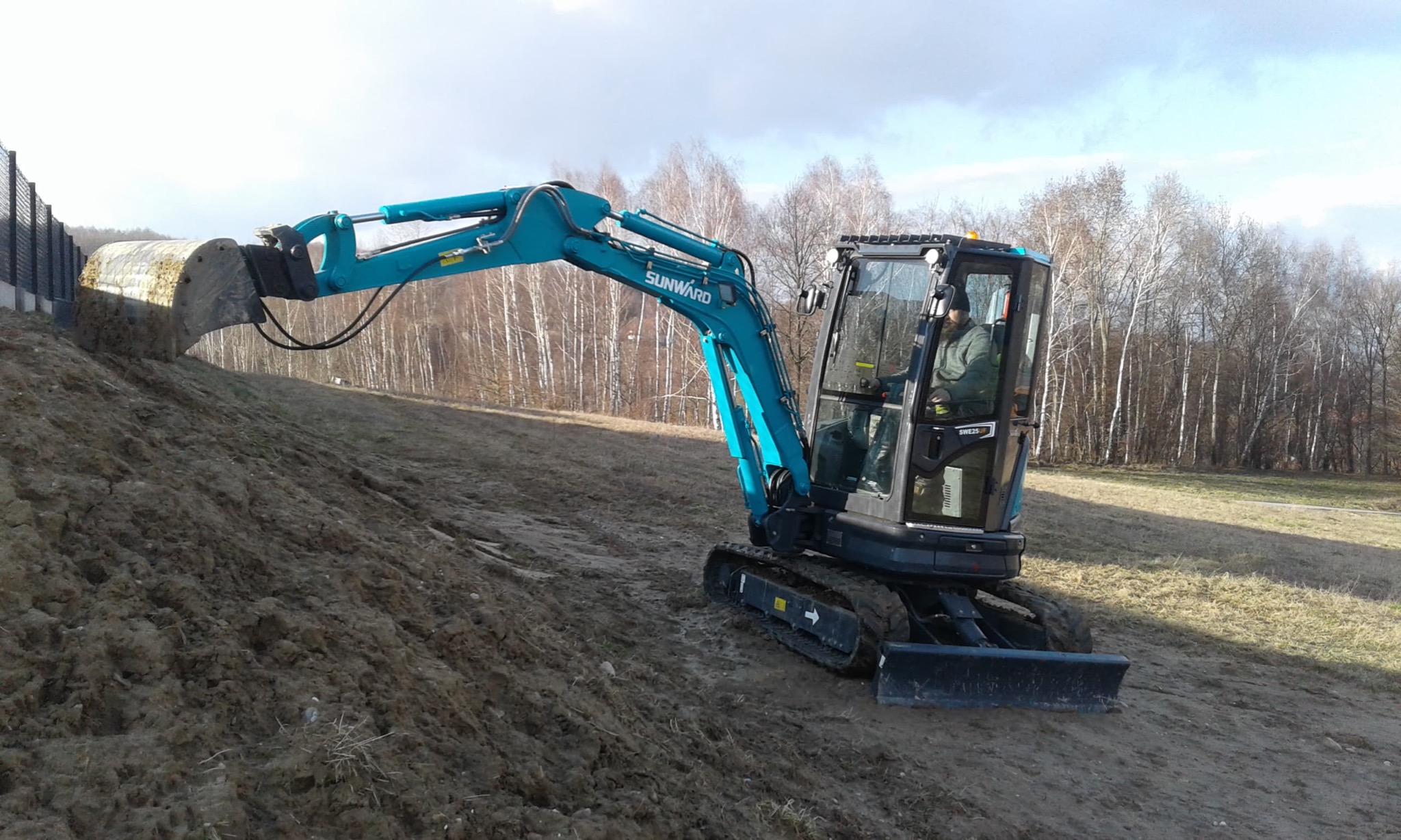 Niebieska mini koparka Sunward SWE25 pracuje na zboczu ziemnym, z operatorem w kabinie, w tle drzewa bez liści i fragment ogrodzenia.