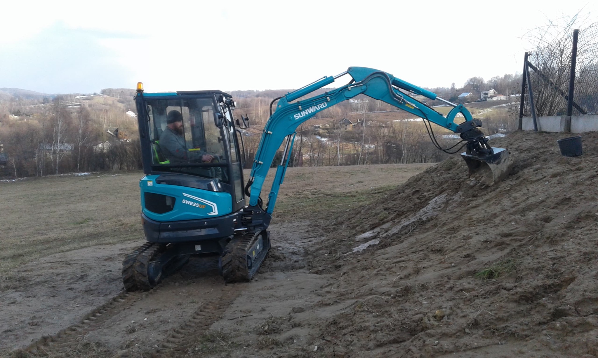 Niebieska mini koparka Sunward SWE25UF z operatorem w kabinie pracuje przy pryzmie ziemi na tle pagórkowatego krajobrazu.