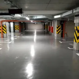 Błyszcząca, szara posadzka w garażu podziemnym z żółto-czarnymi pasami ostrzegawczymi na filarach i wyraźnymi liniami parkingowymi.