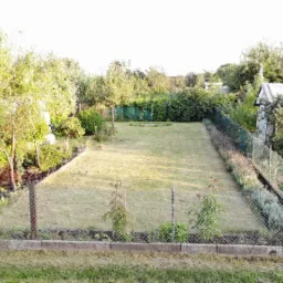 Widok z góry na wypielęgnowany trawnik otoczony drzewami, krzewami i szklarnią, ogrodzony siatką z betonową podmurówką.