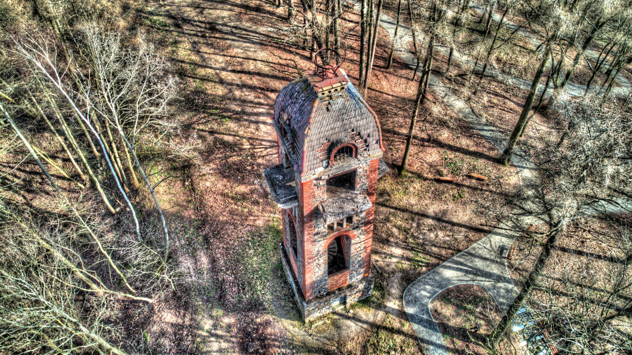 Widok z góry na opuszczoną, ceglaną wieżę z zniszczonym dachem, otoczoną drzewami w parku, z widoczną ścieżką spacerową.