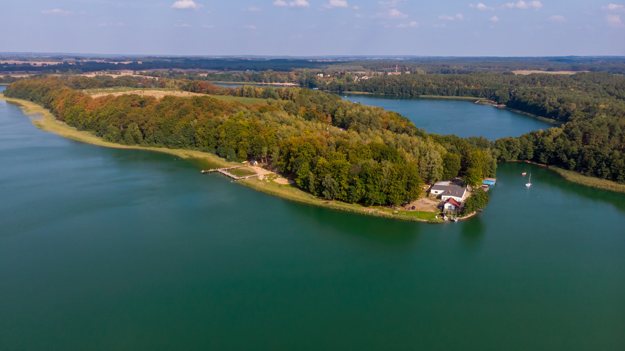 Panoramiczny widok z lotu ptaka na jezioro otoczone lasami, z widoczną przystanią i budynkami na brzegu, uchwycony w słoneczny dzień.