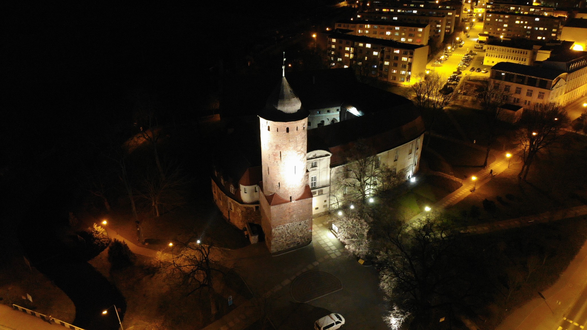 Nocny widok z lotu ptaka na oświetlony zamek z ceglaną wieżą i stożkowym dachem, otoczony budynkami mieszkalnymi i parkiem z latarniami.