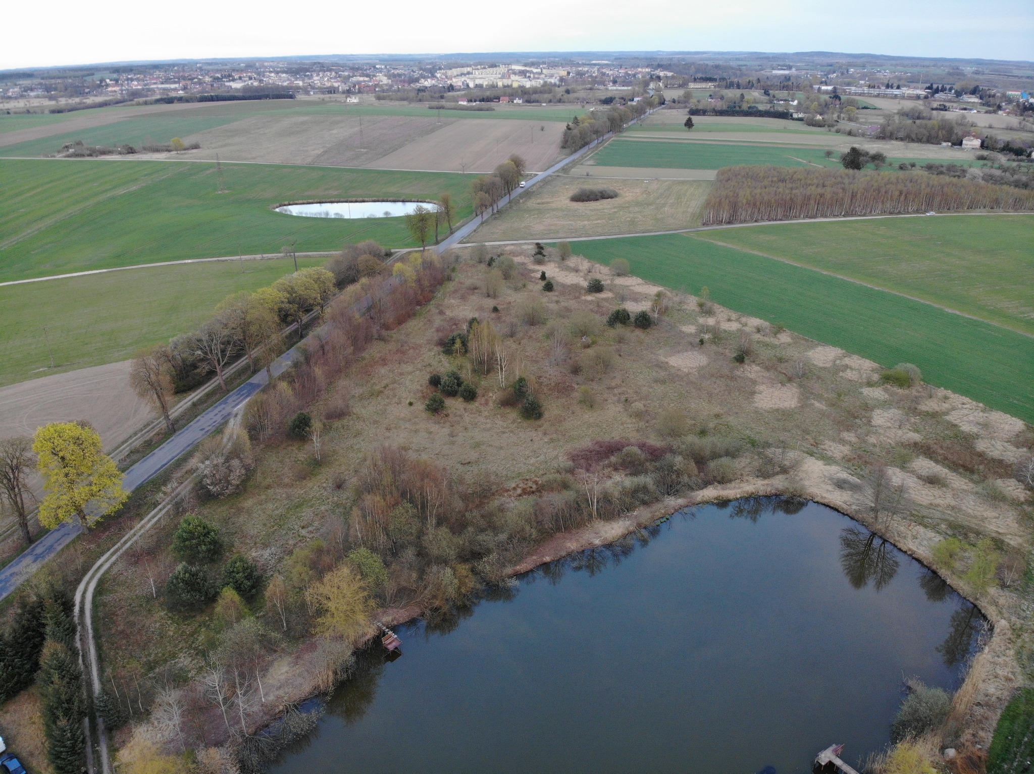 Widok z drona na krajobraz rolniczy z polami, drogą, małym jeziorem i zabudowaniami w oddali, w pochmurny dzień.