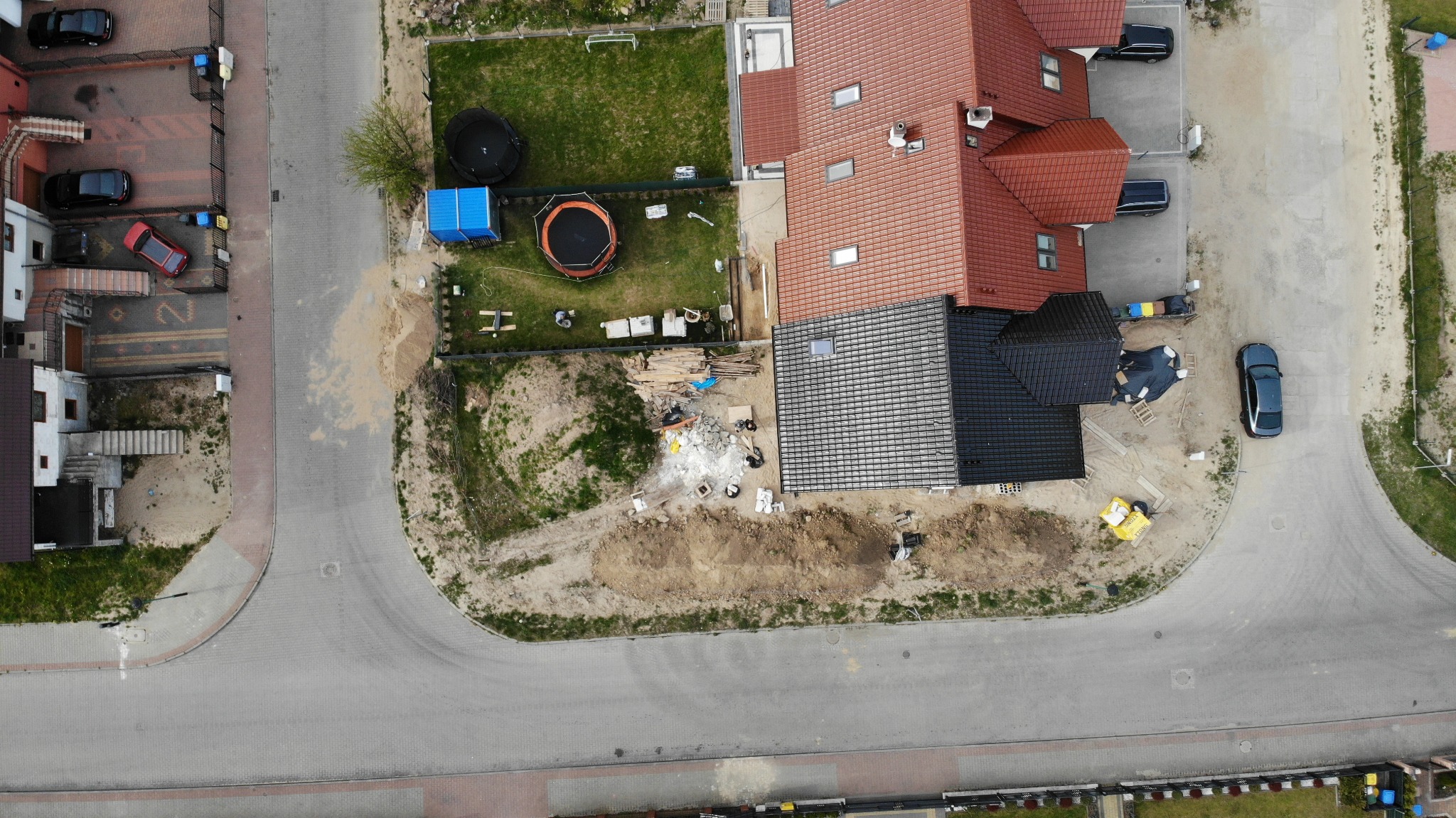 Zdjęcie z drona przedstawia budowę domu jednorodzinnego z czarną dachówką, obok stoi dom z czerwoną dachówką, na podwórku trampoliny, plac budowy z materiałami budowlanymi i wykopaną ziemią.