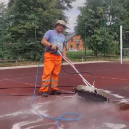 Pracownik w pomarańczowych spodniach i kapeluszu czyści czerwoną nawierzchnię boiska sportowego za pomocą specjalistycznej maszyny ciśnieniowej, w tle siatka i zieleń drzew.