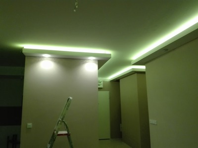 Wykończone wnętrze z podwieszanym sufitem z oświetleniem LED, widoczna drabina malarska, ściany w odcieniach beżu, oświetlenie punktowe.