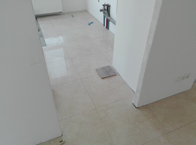 Ułożone beżowe płytki podłogowe w pomieszczeniu w trakcie prac wykończeniowych, widoczne fragmenty instalacji hydraulicznej i płytka testowa na podłodze