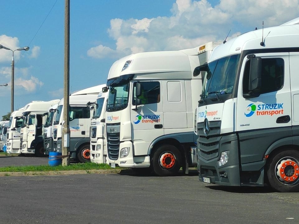 Flota białych ciężarówek firmy Strazik Transport zaparkowana w rzędzie, widoczny DAF i Mercedes Actros, w tle błękitne niebo z chmurami.