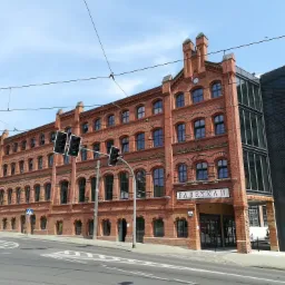 Odrestaurowany, ceglany budynek dawnej fabryki z nowoczesną dobud&oacute;wką, widoczny z poziomu ulicy z sygnalizacją świetlną.