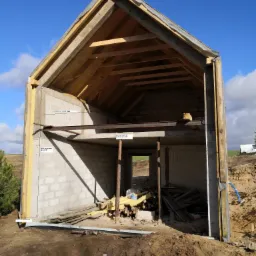 Surowy stan budynku w trakcie budowy; widoczne ściany z bloczków betonowych, drewniana konstrukcja dachu, stalowe belki nośne, izolacja termiczna na zewnętrznej ścianie oraz ziemia wokół fundamentów.