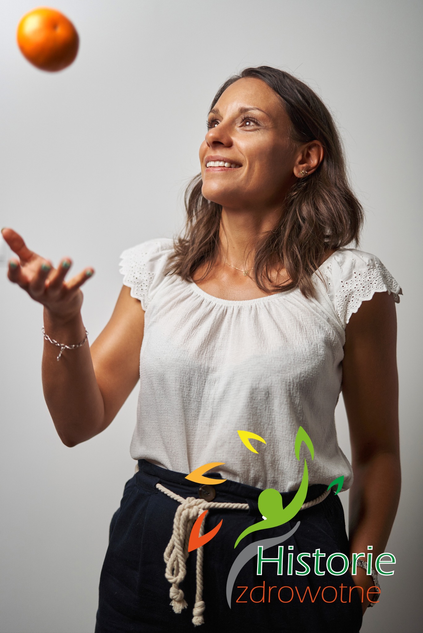 Uśmiechnięta kobieta w białej bluzce i granatowych spodniach, z mandarynką unoszącą się w powietrzu nad jej dłonią, na dole widoczne logo 'Historie zdrowotne'.