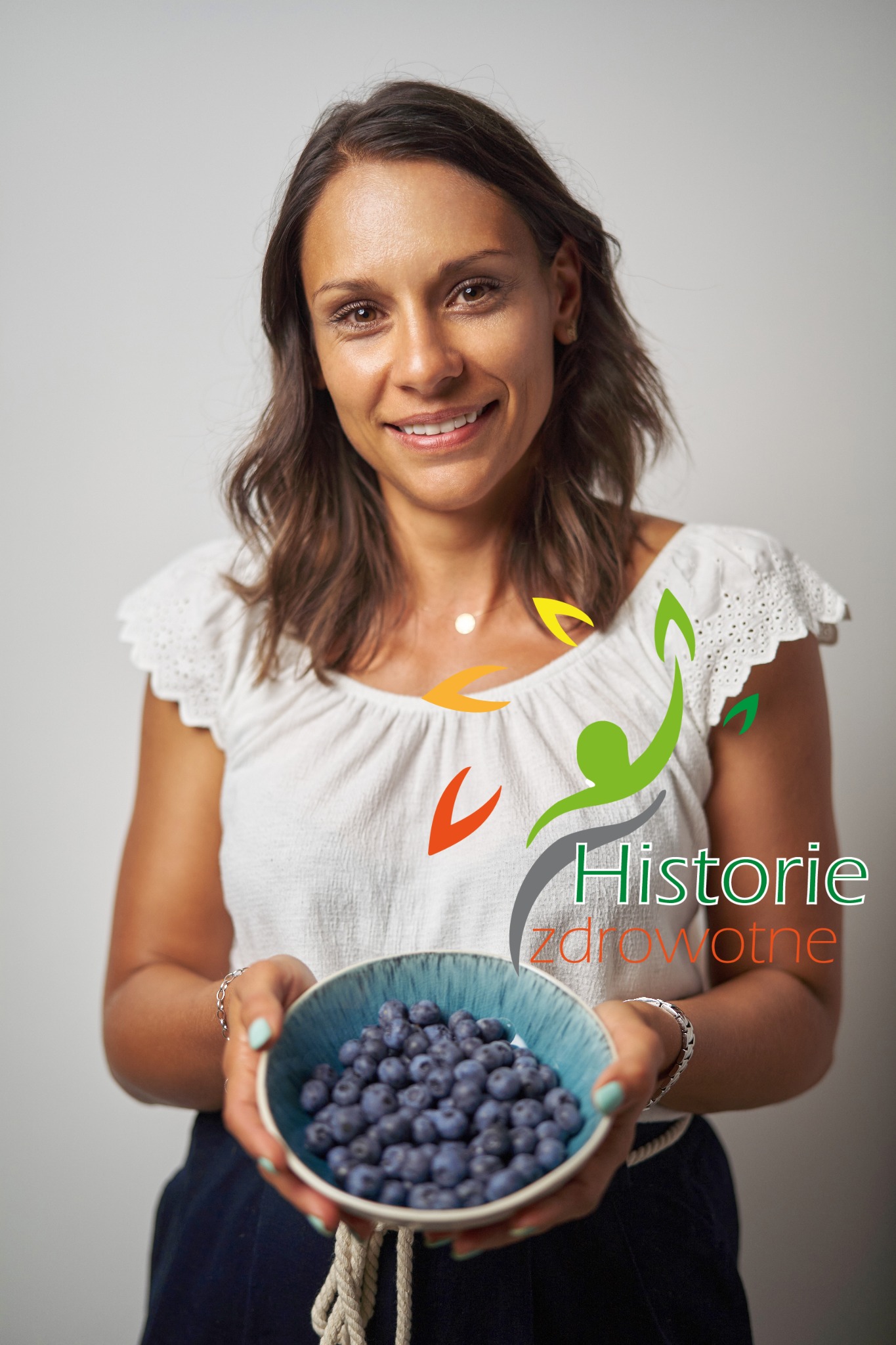Uśmiechnięta kobieta trzymająca miseczkę pełną borówek, z logo 'Historie Zdrowotne' nałożonym na zdjęcie.