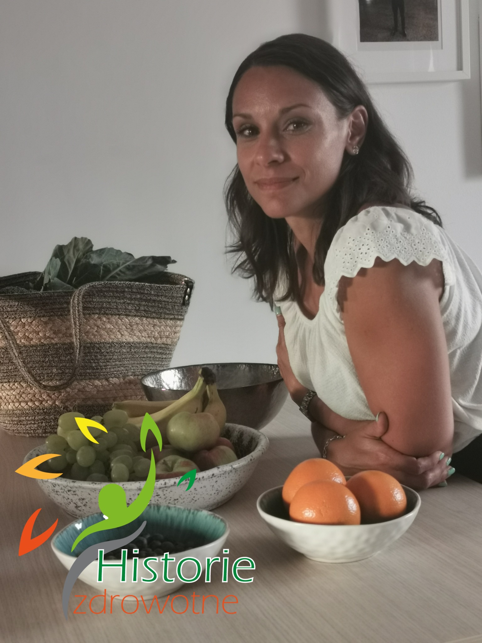 Uśmiechnięta kobieta opiera się o stół z misami pełnymi owoców: winogron, bananów, jabłek, pomarańczy i jagód; w tle torba z zielonymi liśćmi warzyw.