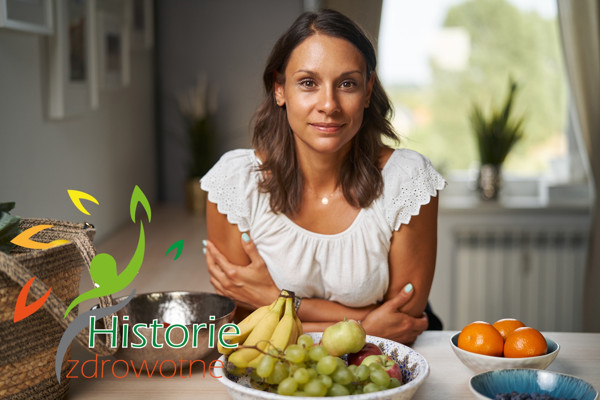 Kobieta z brązowymi włosami i w białej bluzce siedzi za stołem z miskami owoców: bananami, winogronami, jabłkami, pomarańczami i jagodami. W lewym dolnym rogu widoczne logo 'Historie zdrowotne'.