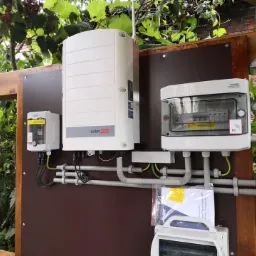 Zewnętrzna instalacja komponentów systemu zasilania energią słoneczną, w tym falownik SolarEdge, zabezpieczenia i skrzynka przyłączeniowa, zamontowane na brązowej płycie na drewnianej ramie,...