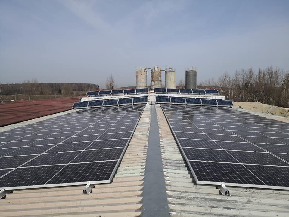 Rozległa instalacja fotowoltaiczna na dachu przemysłowym z widocznymi silosami w tle, uchwycona w pochmurny dzień z perspektywy lekko z góry.