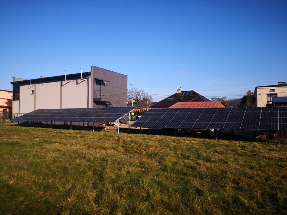 Dwie rzędy paneli słonecznych na metalowych konstrukcjach, ustawione na trawiastym terenie przed budynkami w słoneczny dzień.