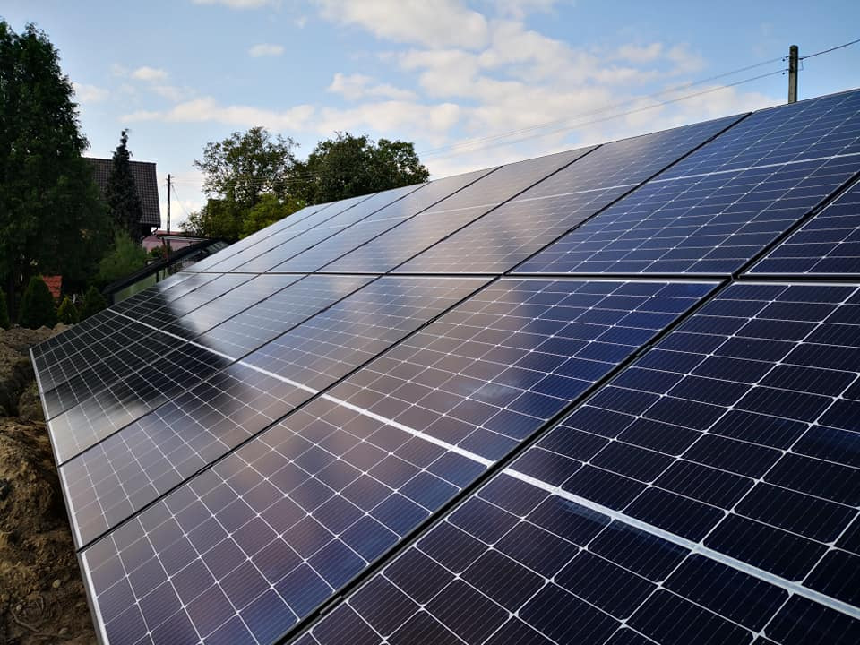 Panel słoneczny z widocznym odbiciem nieba, zainstalowany na dachu, z ziemią widoczną u podstawy instalacji.