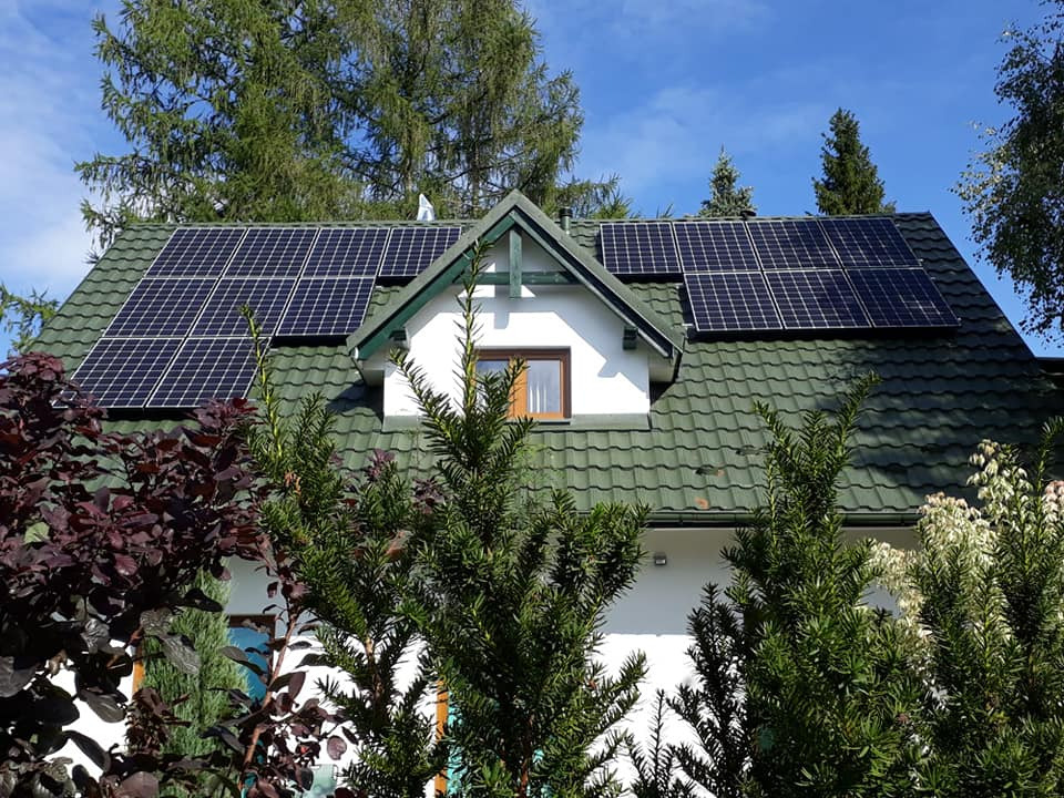 Panele słoneczne na zielonym dachu domu jednorodzinnego, widoczne zza gęstych krzewów w słoneczny dzień.