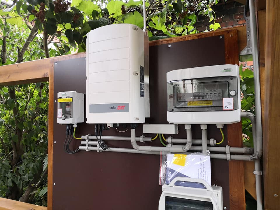 Zewnętrzna instalacja komponentów systemu zasilania energią słoneczną, w tym falownik SolarEdge, zabezpieczenia i skrzynka przyłączeniowa, zamontowane na brązowej płycie na drewnianej ramie,...