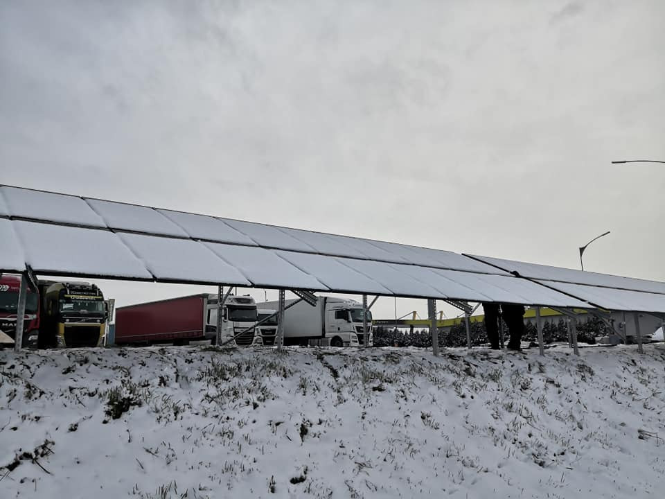 Rząd paneli słonecznych pokrytych śniegiem, ustawionych jako zadaszenie nad parkingiem dla ciężarówek w zimowy dzień.