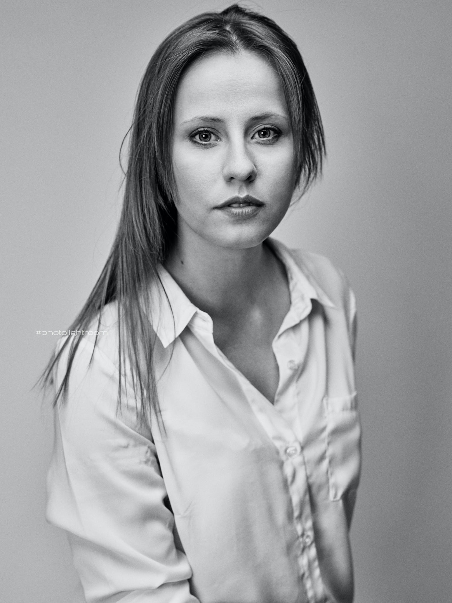 Czarno-białe portretowe ujęcie kobiety w białej koszuli, z długimi prostymi włosami, skierowanej wzrokiem w obiektyw na jednolitym szarym tle.