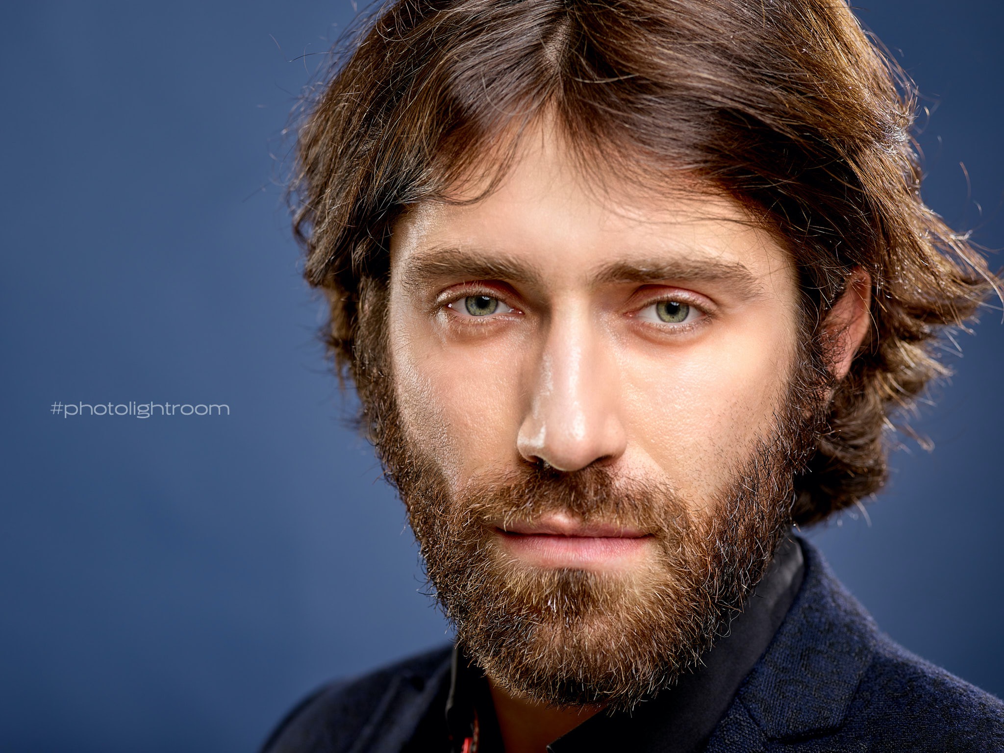 Portret brodatego mężczyzny z zielonymi oczami na ciemnoniebieskim tle, z widocznym hashtagiem #photolightroom.