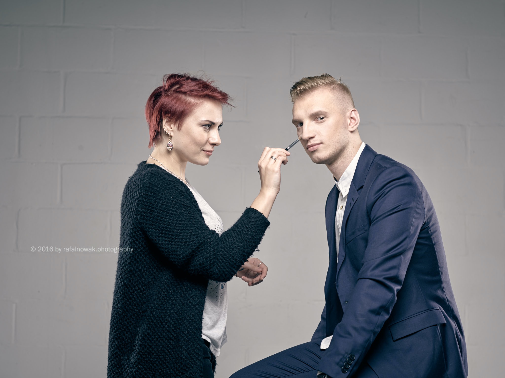 Wizażystka poprawia makijaż eleganckiego mężczyzny w granatowym garniturze, przygotowując go do sesji zdjęciowej na tle białej ściany z cegieł.