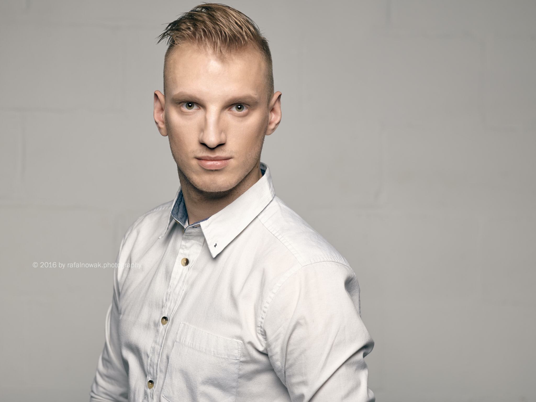 Portret młodego mężczyzny w białej koszuli, z charakterystyczną fryzurą i poważnym spojrzeniem, na jasnoszarym tle.