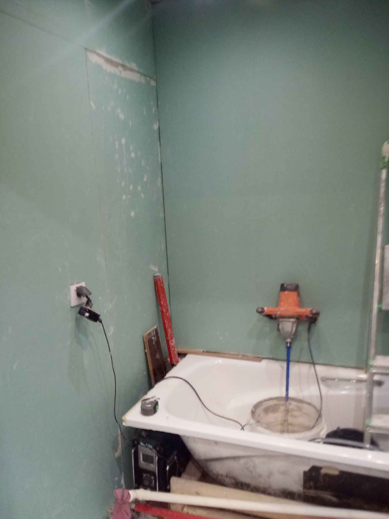 Niedokończona łazienka z zielonymi ścianami gipsowymi, białą wanną wypełnioną wiadrem z zaprawą, mieszadłem elektrycznym i narzędziami budowlanymi w nieładzie.