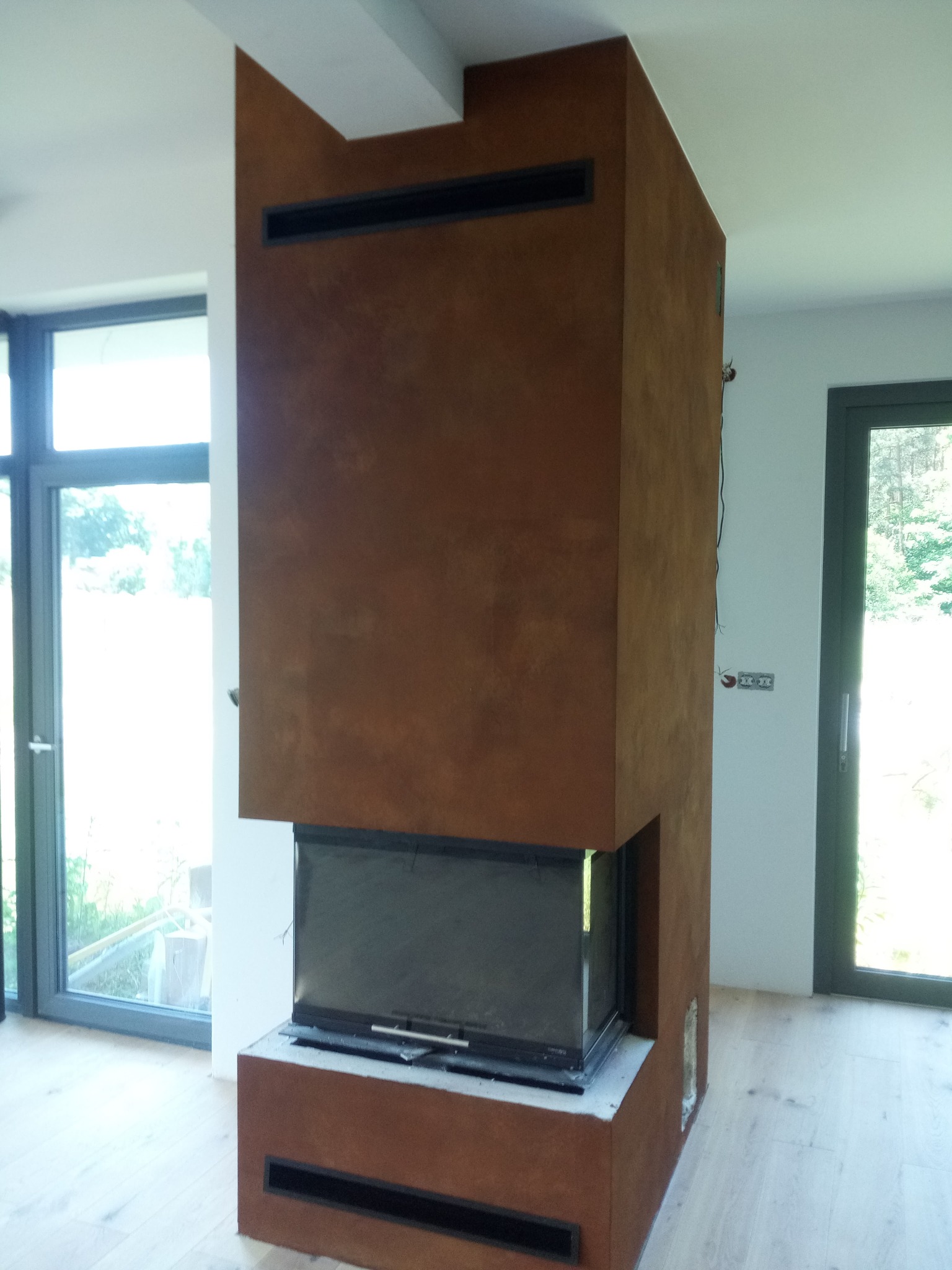 Nowoczesny kominek obudowany płytami imitującymi rdzę, z czarnymi wlotami powietrza, w jasnym wnętrzu z dużymi oknami i drewnianą podłogą.