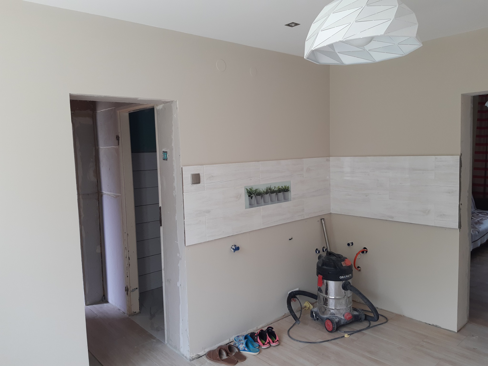 Fragment wnętrza w trakcie remontu: częściowo położone płytki imitujące drewno nad blatem, przygotowane punkty instalacji wodnej, odkurzacz przemysłowy i buty robocze na podłodze, surowa ściana...