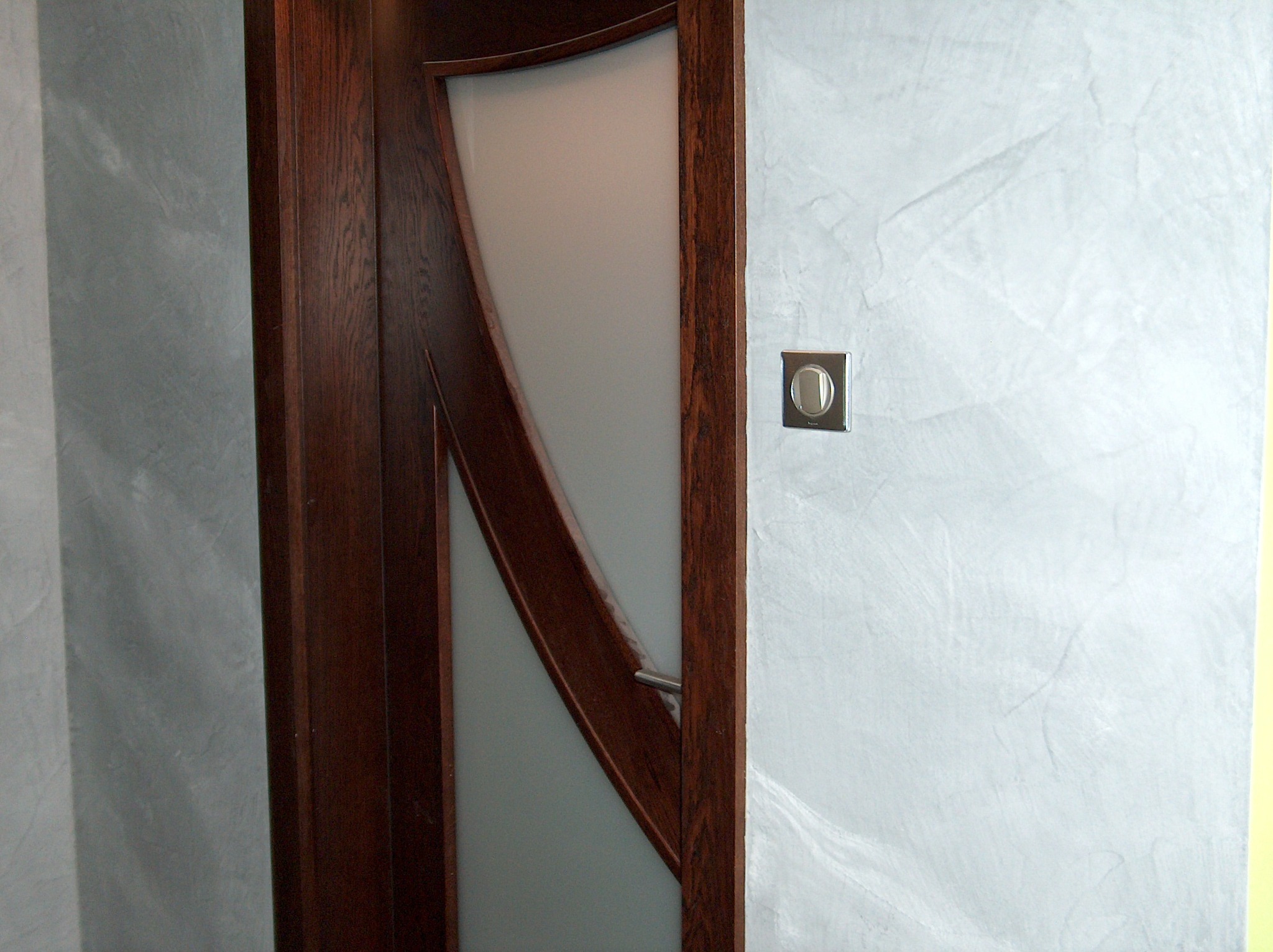 Ściana z efektem dekoracyjnym tynku marmorino w odcieniach szarości, obok drewniane drzwi z matową szybą i chromowanym uchwytem oraz włącznik światła.