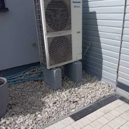 Biała jednostka zewnętrzna klimatyzacji Panasonic Inverter, umieszczona na betonowych bloczkach, otoczona kamiennym grysem i kostką brukową przy szarej ścianie budynku.