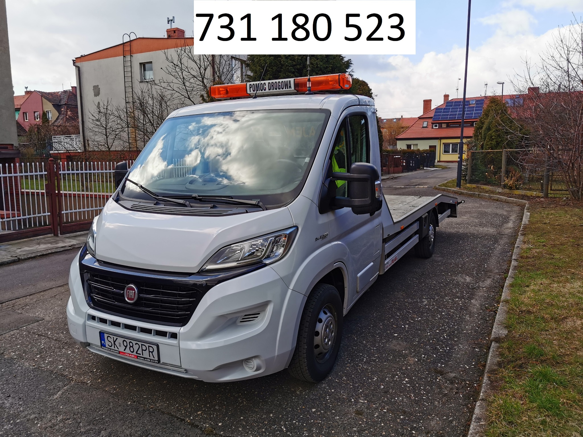 Biała laweta Fiat z pomarańczowym świetłem ostrzegawczym 'Pomoc Drogowa' na dachu, zaparkowana na osiedlowej uliczce.