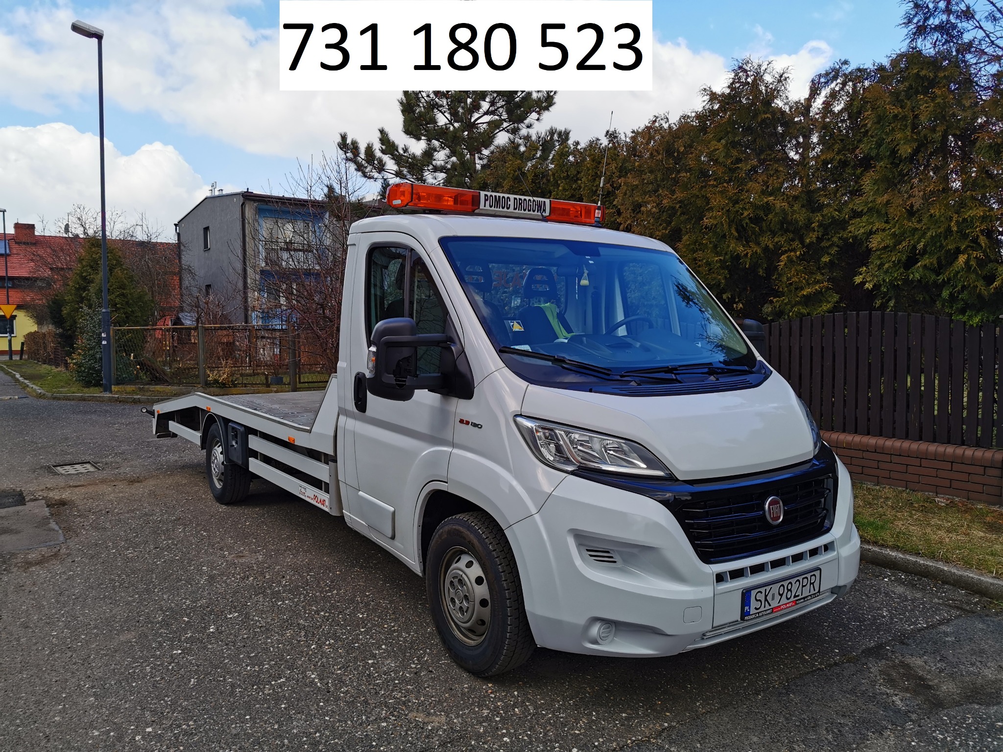 Biała laweta Fiat Ducato z napisem 'Pomoc Drogowa' na pomarańczowym świetlnym panelu, zaparkowana na asfaltowej drodze w pobliżu budynku i ogrodzenia.