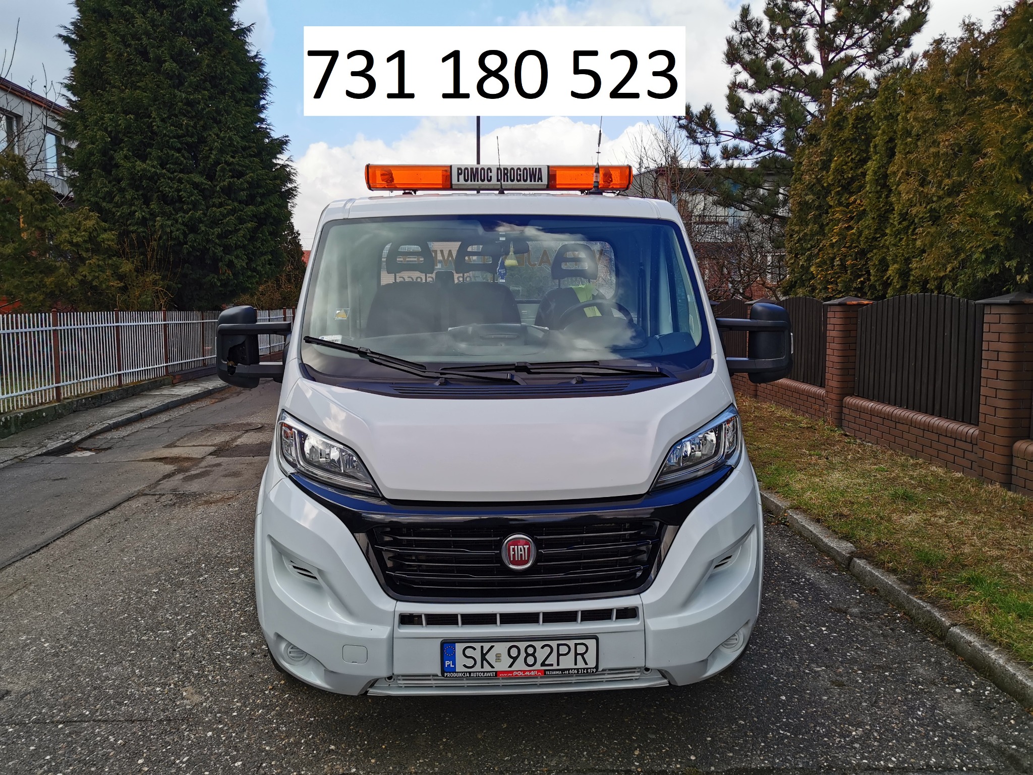 Biały Fiat Ducato z napisem 'Pomoc Drogowa' i numerem telefonu na szybie, zaparkowany na ulicy.