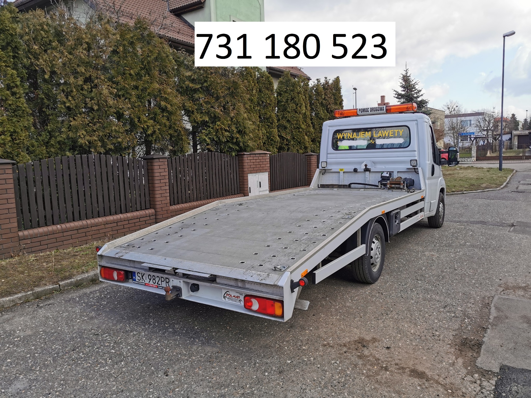 Biała laweta z napisem 'Wynajem Lawety' widziana z tyłu na tle zabudowań i zieleni, z numerem telefonu na zdjęciu.